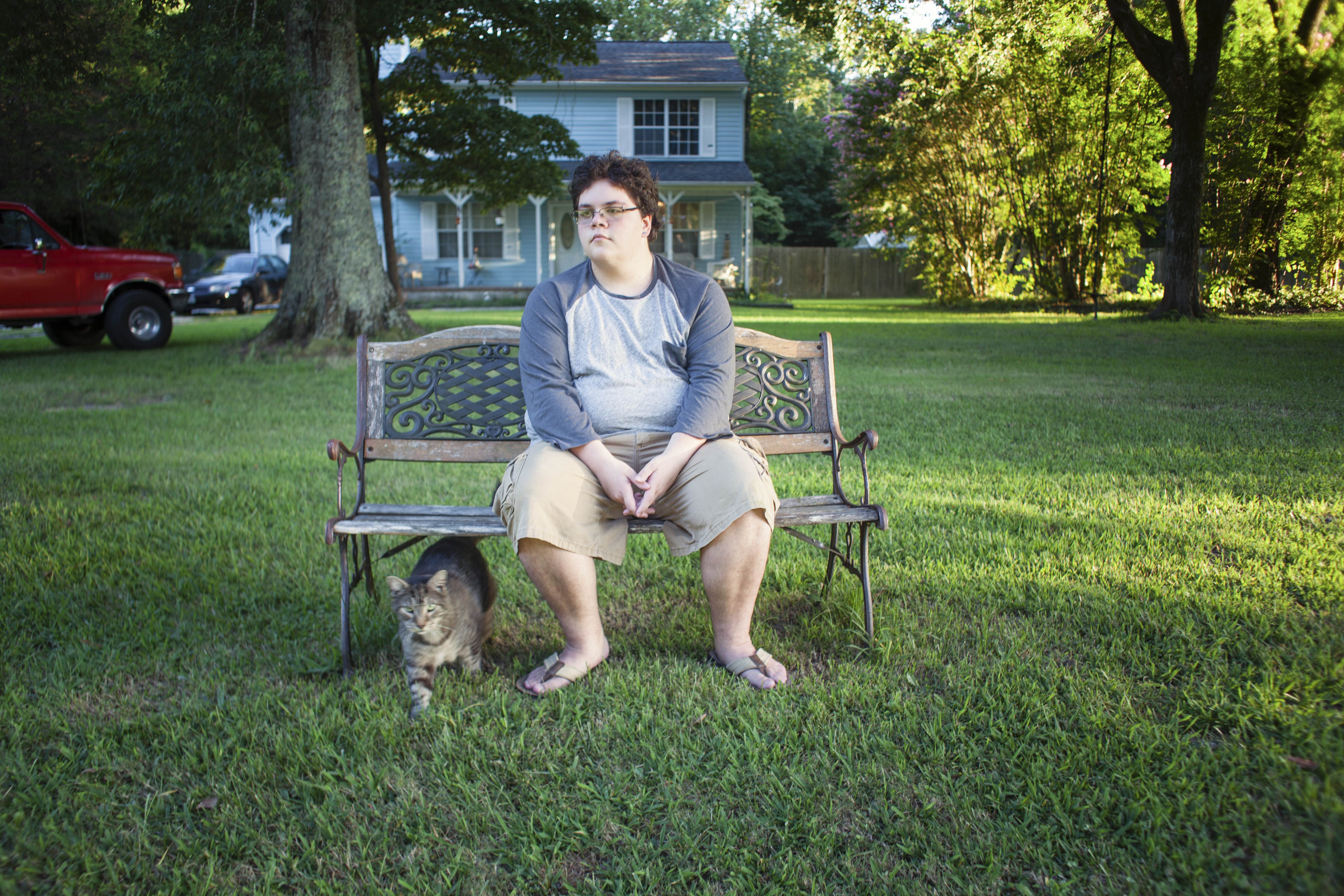 Gavin Grimm sits on a bench with a cat by his feet and a house and truck in the background.