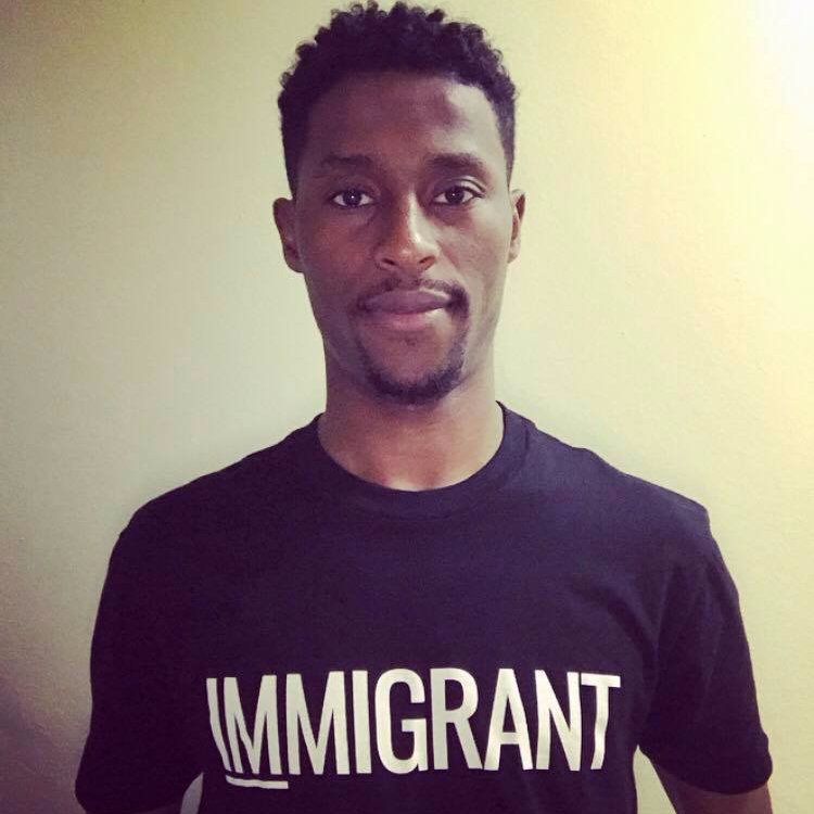A black male standing against a white background, wearing a black shirt with white writing saying "Immigrant"
