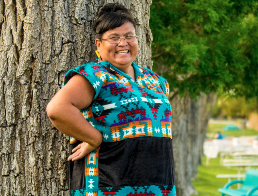 Diné trans leader Mattee Jim stands outside, back against a tree, smiling