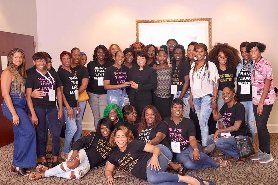 Group of 30 Black transgender people, most in Black Trans Lives Matter shirts, smile and hug
