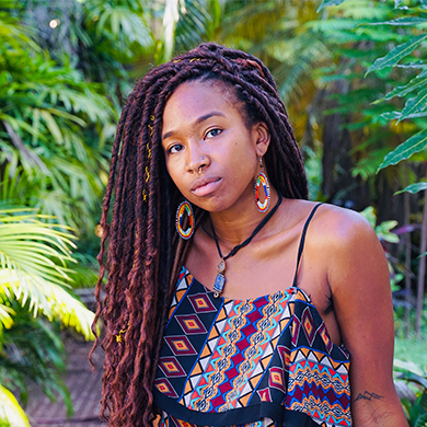 A black person, wearing African pattern earings and clothing, They have long dreads.