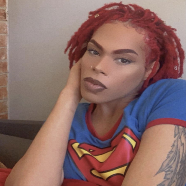 Phoenix Gayle, a Black trans woman, wearing a Superman t-shirt. She has chin-length dyed red hair and an upper arm tattoo.