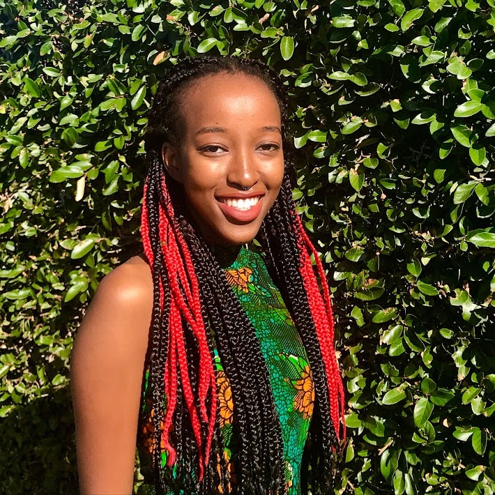 Karīmi Ndwiga, a Black person with long brown and bright red hair, their body angled slightly to the right while smiling at the camera. She stands in front of a tall hedge wearing a green patterned blouse.