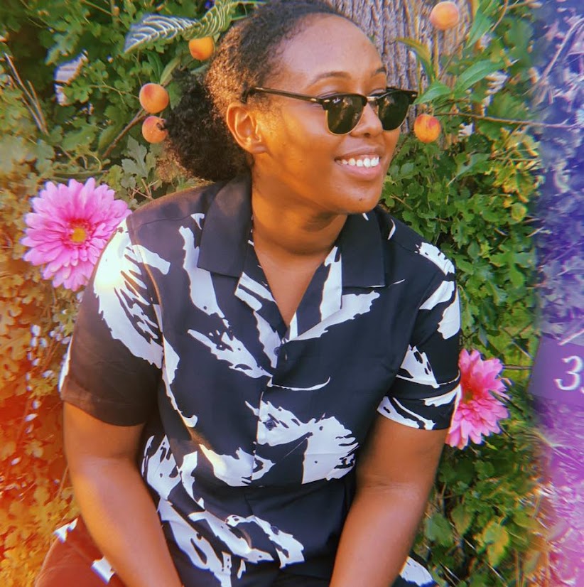 A Black woman with her hair tied back, glancing slightly off to the right. She wears Ray-Ban sunglasses and a black button-down shirt with white leaves. A bush in the background bears a pink flower and small peaches. The margins of the image have an orange-y film camera glow.