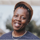 NolizweNondabula A Black person with short dark hair, grinning wide with their head held at an angle. They wear a brown short-brimmed cap, a blue t-shirt, and a silver metal necklace.