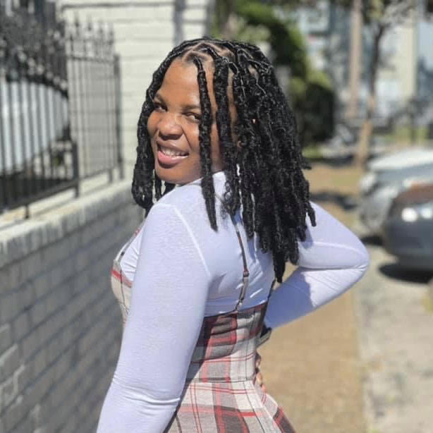 Aneiry L. Zapata, a Black person with long dark hair, with their back turned to the camera. They're looking over their shoulder and smiling, standing on a sidewalk near a row of cars, wearing a light purple long-sleeve blouse and red and white checkered coveralls.