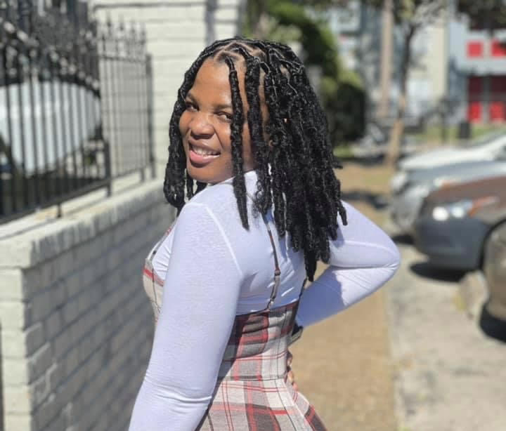 Aneiry L. Zapata, a Black person with long dark hair, with their back turned to the camera. They're looking over their shoulder and smiling, standing on a sidewalk near a row of cars, wearing a light purple long-sleeve blouse and red and white checkered coveralls.