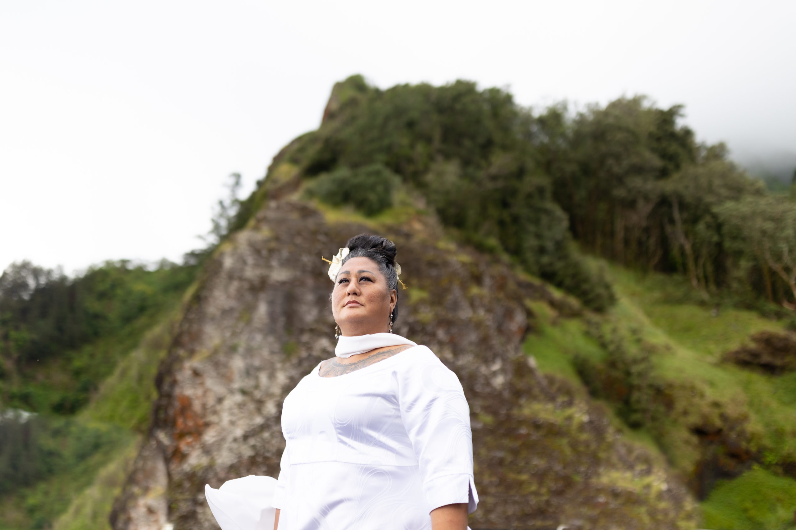 A photo of Kumu Hina, a brown-skinned trans woman and mahu, wearing a white dress with a silk scarf and a matching white flower in her hair.
