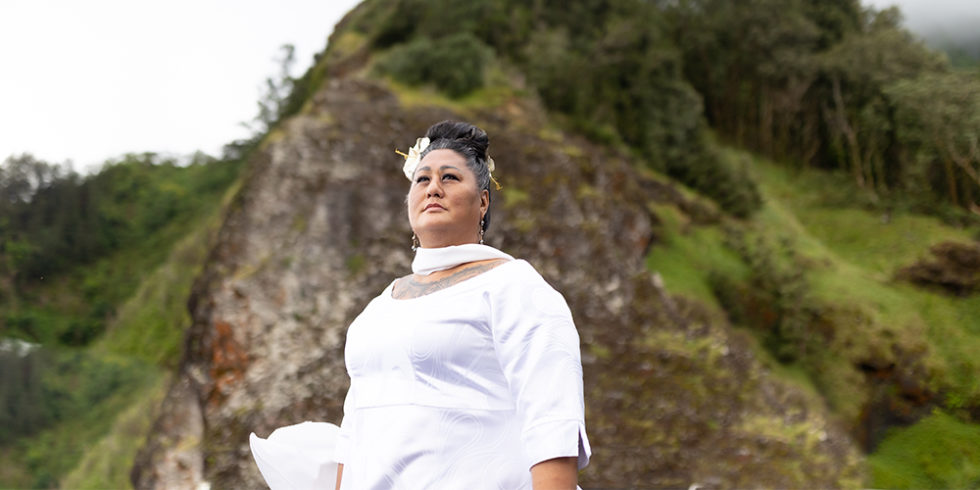 A brown-skinned woman wearing a white gown and white flower in her hair, standing against a mountain.