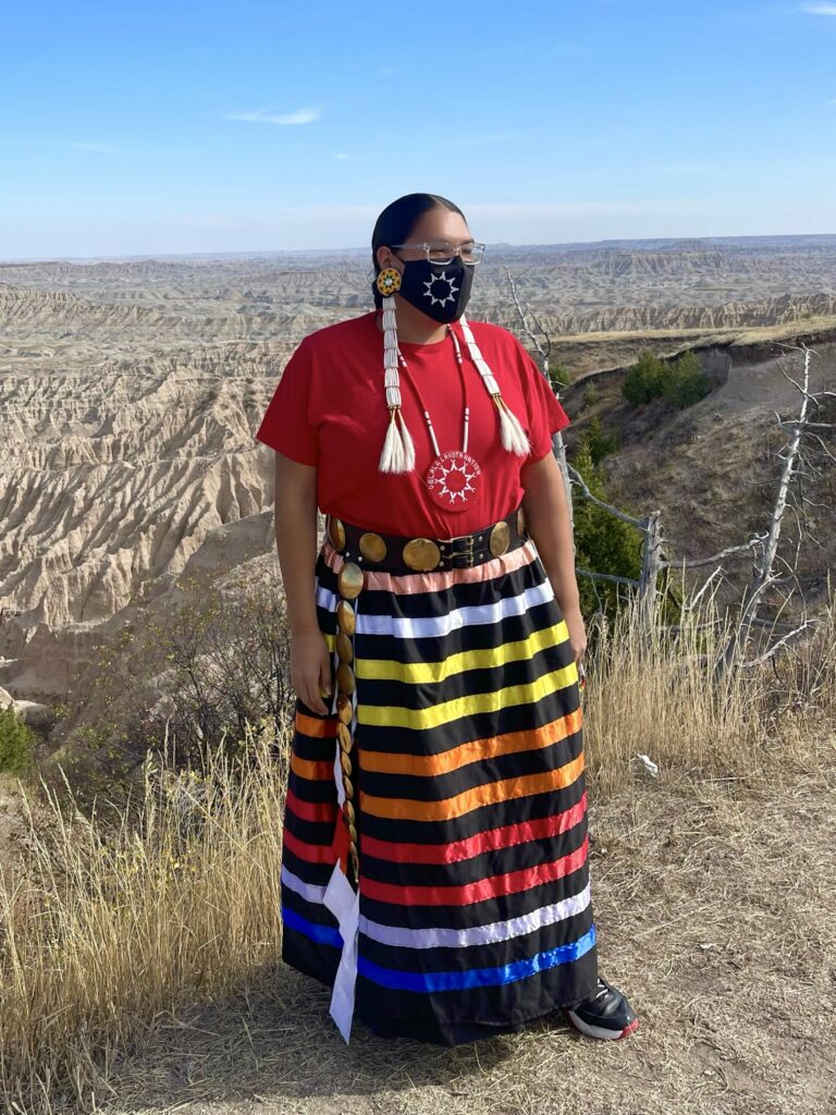 A photo of a brown-skinned person standing at a mountaintop. They wear braids, a red shirt, and a multicolored striped skirt.
