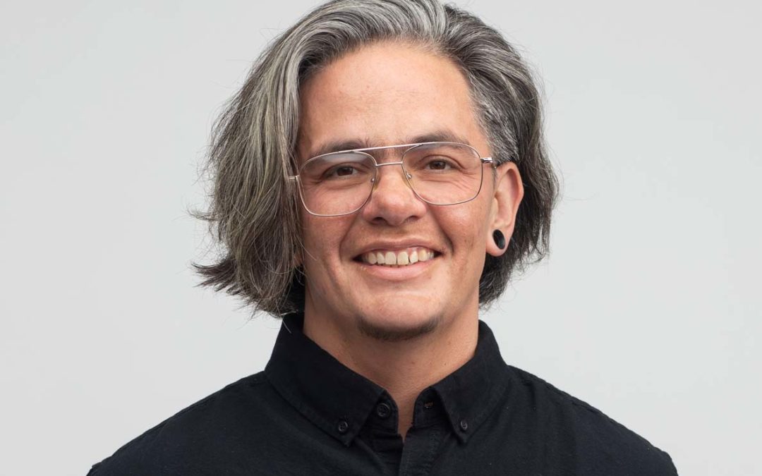 Shelby, a white nonbinary person with chin length gray hair and glasses, smiles at the camera. Their arms are behind their back, and they look confident.