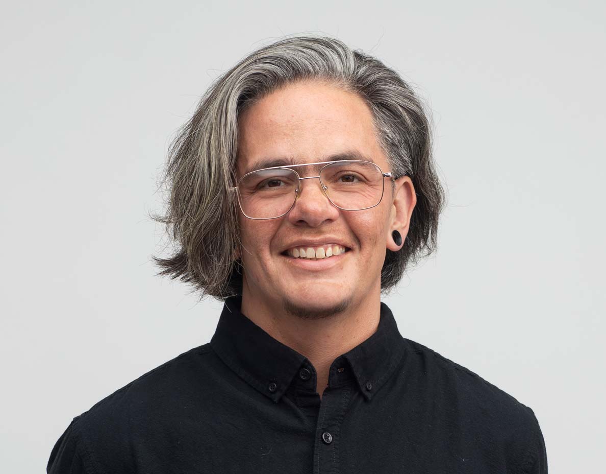 Photo of Shelby Chestnut, a Native American trans person, smiling, with long silver hair and wire glasses, wearing a black shirt.