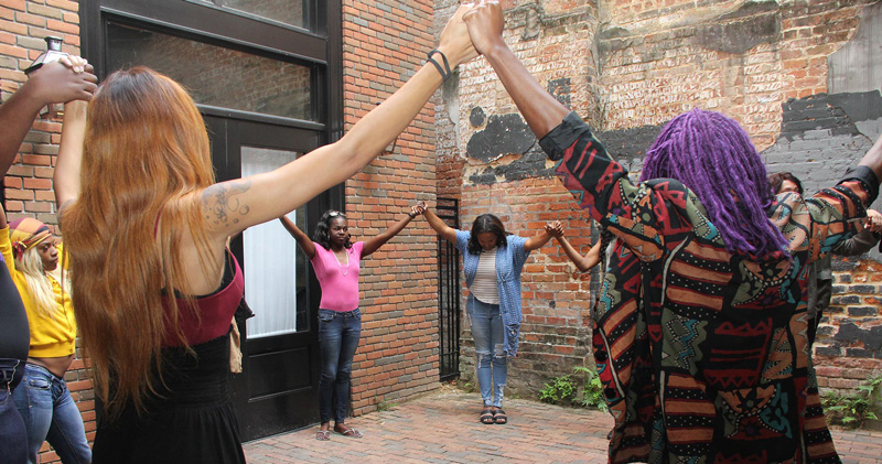 A group of black trans women and femmes standing a circle holding hands and raising them up