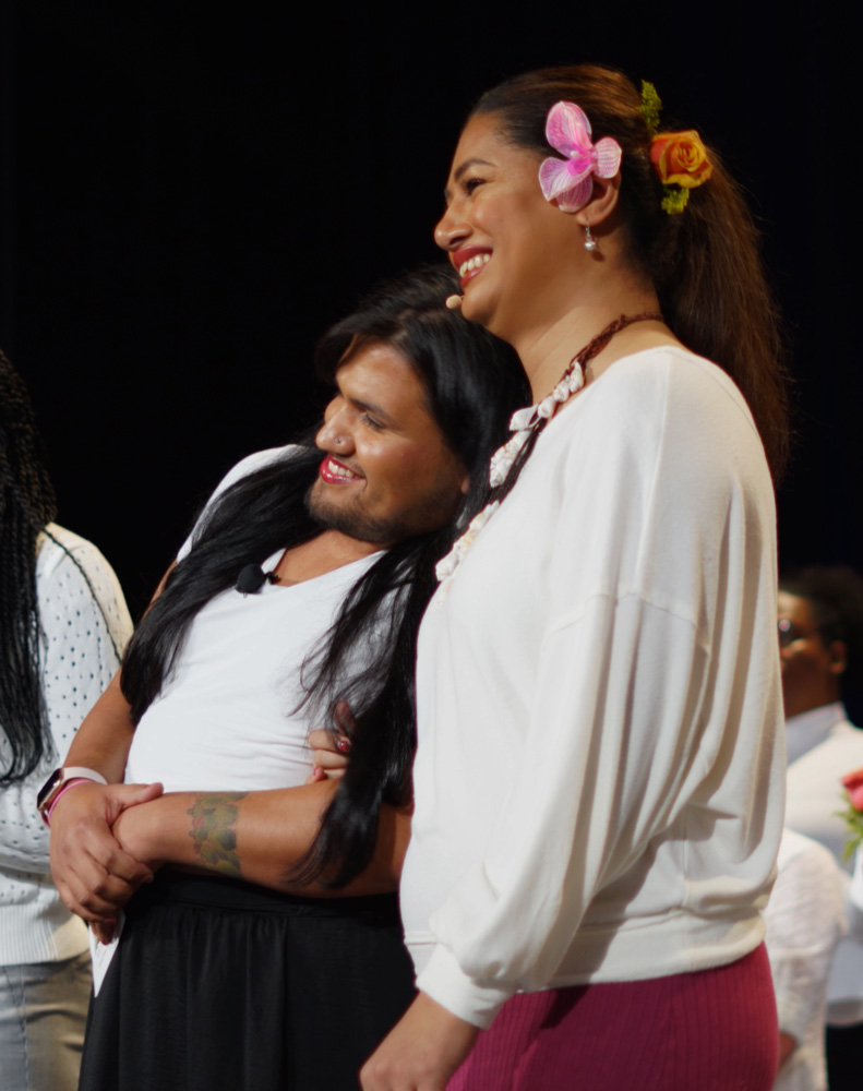 Two people embrace onstage, one leaning into the other. They both have light brown skin and long, dark brown hair. They both wear white shirts.