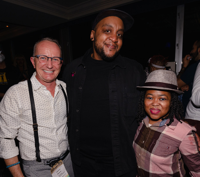 A photo of three people smiling and standing together. Sebastian is a white trans man wearing glasses, a white shirt, and black suspenders. Gabriel is a Black trans man with a goatee, wearing a black hat and black shirt. Ericka is a Black nonbinary femme wearing a gray hat and pink and black checked shirt