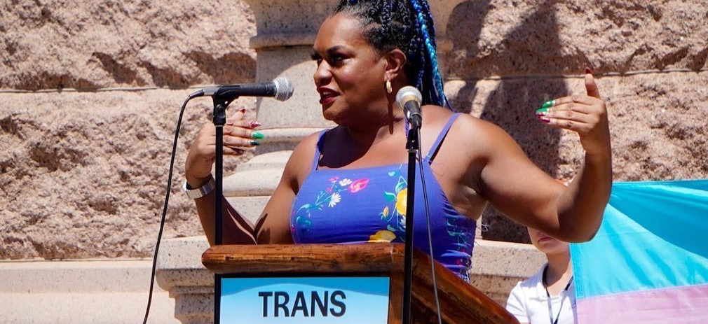 Photo of Imara Jones, a Black trans woman, wearing a blue top with flowers, standing behind a podium, speaking into microphones at a Texas rally.