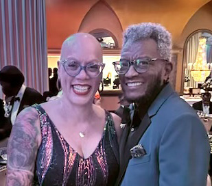 Photo of Tamara, a Black woman with a shaved head, wearing glasses, and sequined dress, and Jay, a Black man with short silver hair, wearing glasses and a blue jacket. 