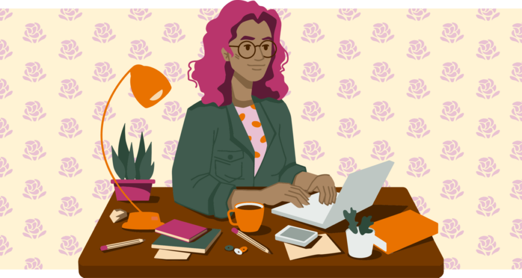 A brown person with wavy pink hair sits at a desk, typing on a small silver laptop. They’re wearing a green jacket with a pocket on the chest, and a pink shirt with orange polka dots. Various items line the desk, including several notebooks, a coffee mug, pencils, buttons, a lamp, and a plant. Behind them is yellow wallpaper with rows of pink decorative roses.