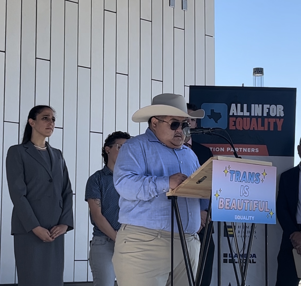 Photo of Emmett, a South Korean American trans man, wearing a beige cowboy hat, dark sunglasses, and blue shirt, standing behind a podium with a Trans is Beautiful sign on it. Lynly is a white cisgender woman, wearing a gray jacket and skirt, standing behind Emmett.