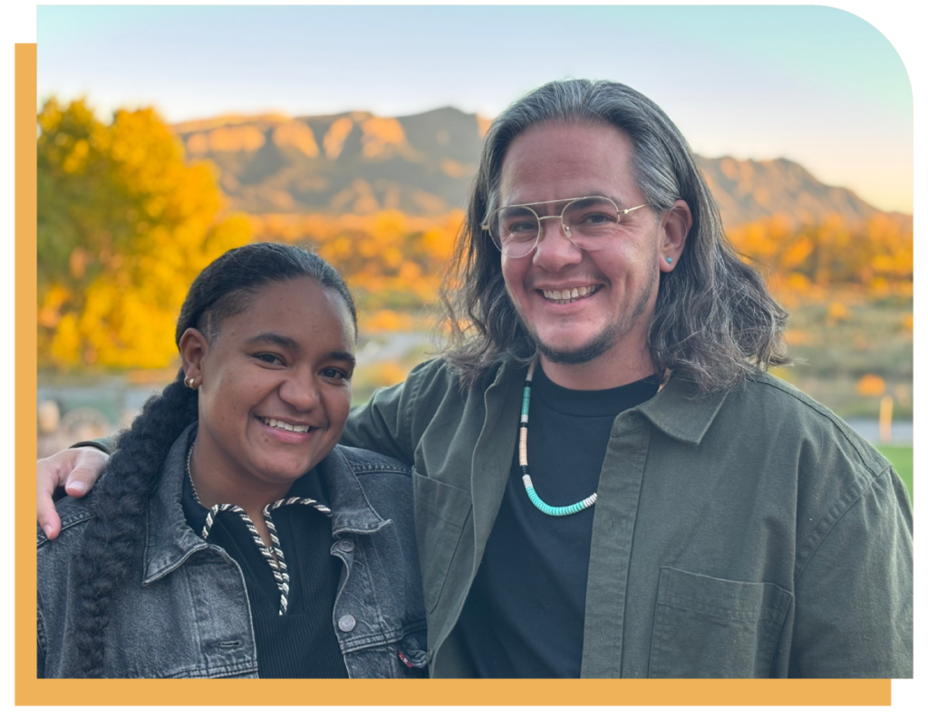 TLC Board Chair Ana Conner, a person with brown skin and braided hair, and TLC Executive Director Shelby Chestnut, a Native American trans person with shoulder length hair and glasses, embrace one another as bold leaders against a background of mountains in golden light