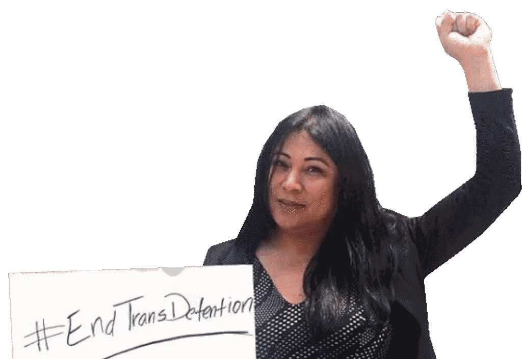 A woman of brown complexion and long black hair is raising a fist and holding a sign that says "End Trans Detention."